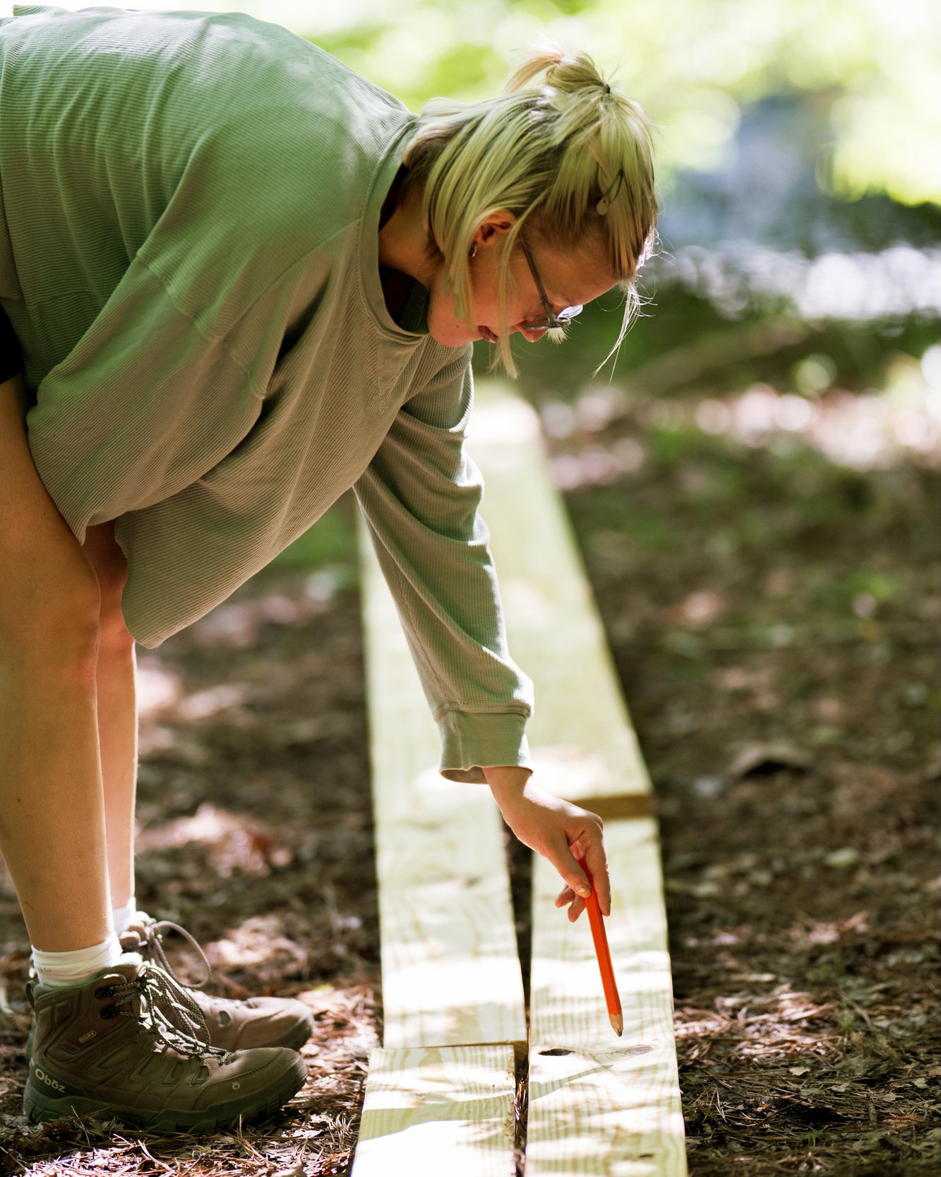 StickBuilt24+(17) Woman leans over a line of lumber as she begins to mark one with a carpenter's pencil