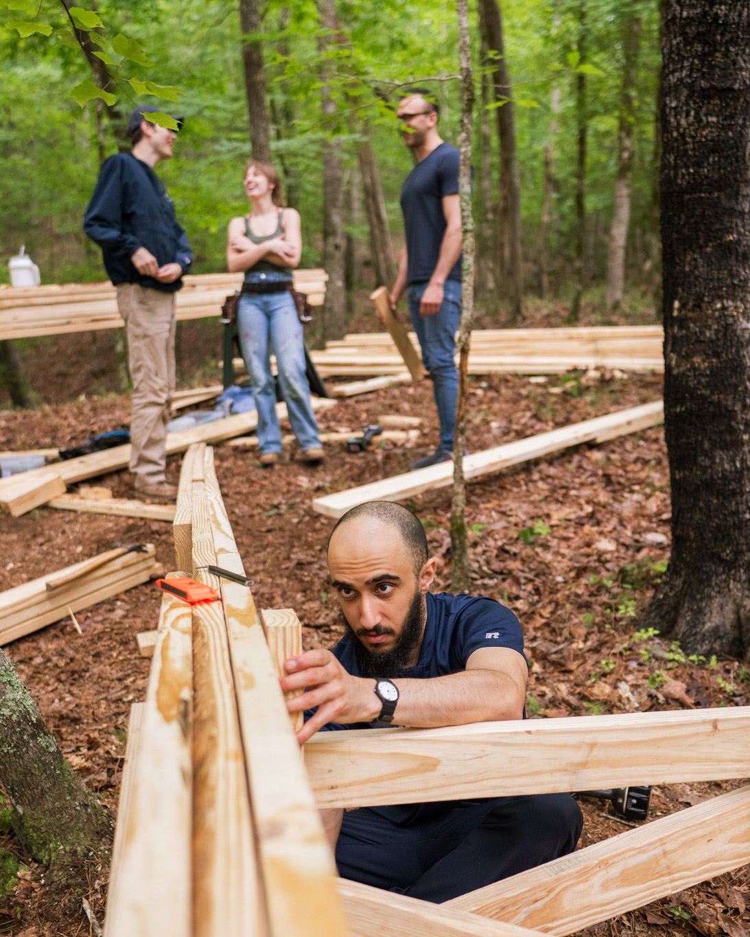 StickBuilt24+(19) Man is leveling some wood, concentrating hard. In the background, a group of three stands in happy conversation, standing among lumber laying on the ground.