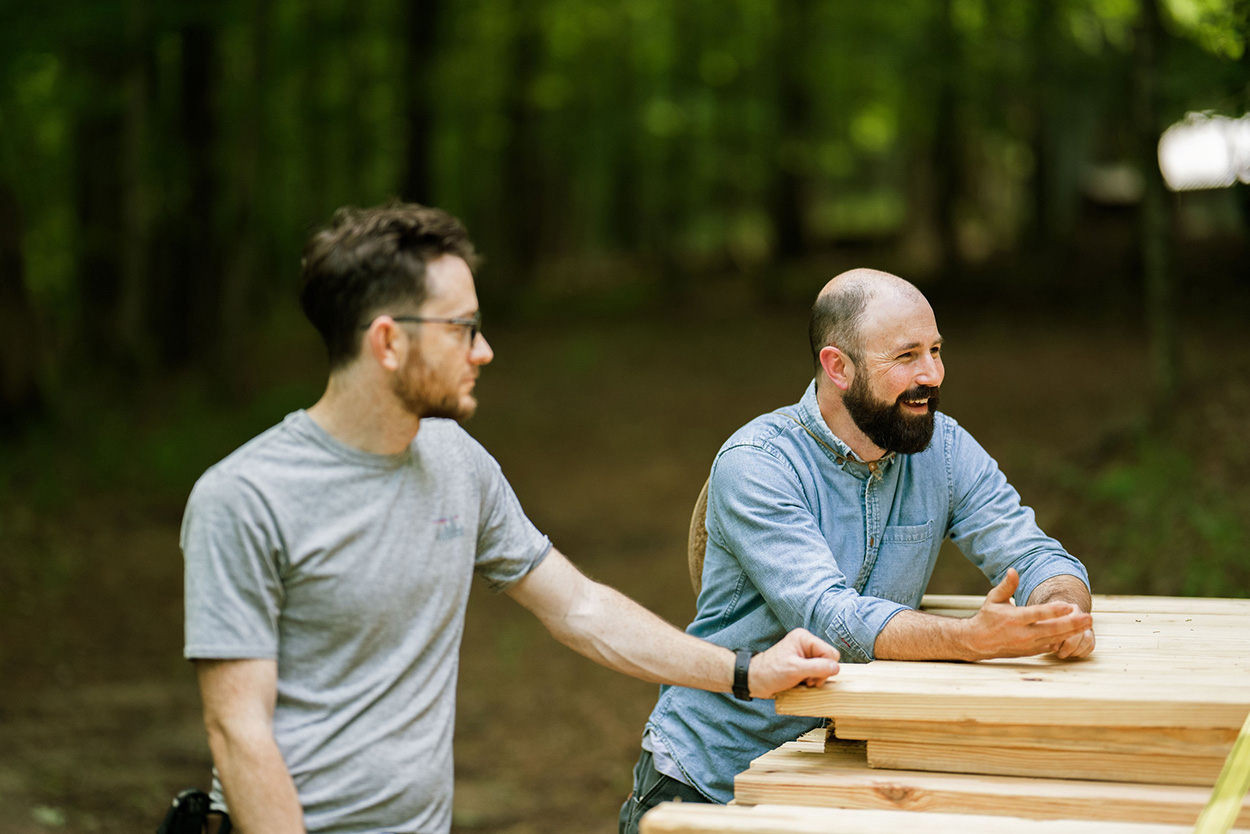 StickBuilt24+(8)@0.5x Will looking in Charlie's direction as Charlie leans on a stack of wood, laughing and talking