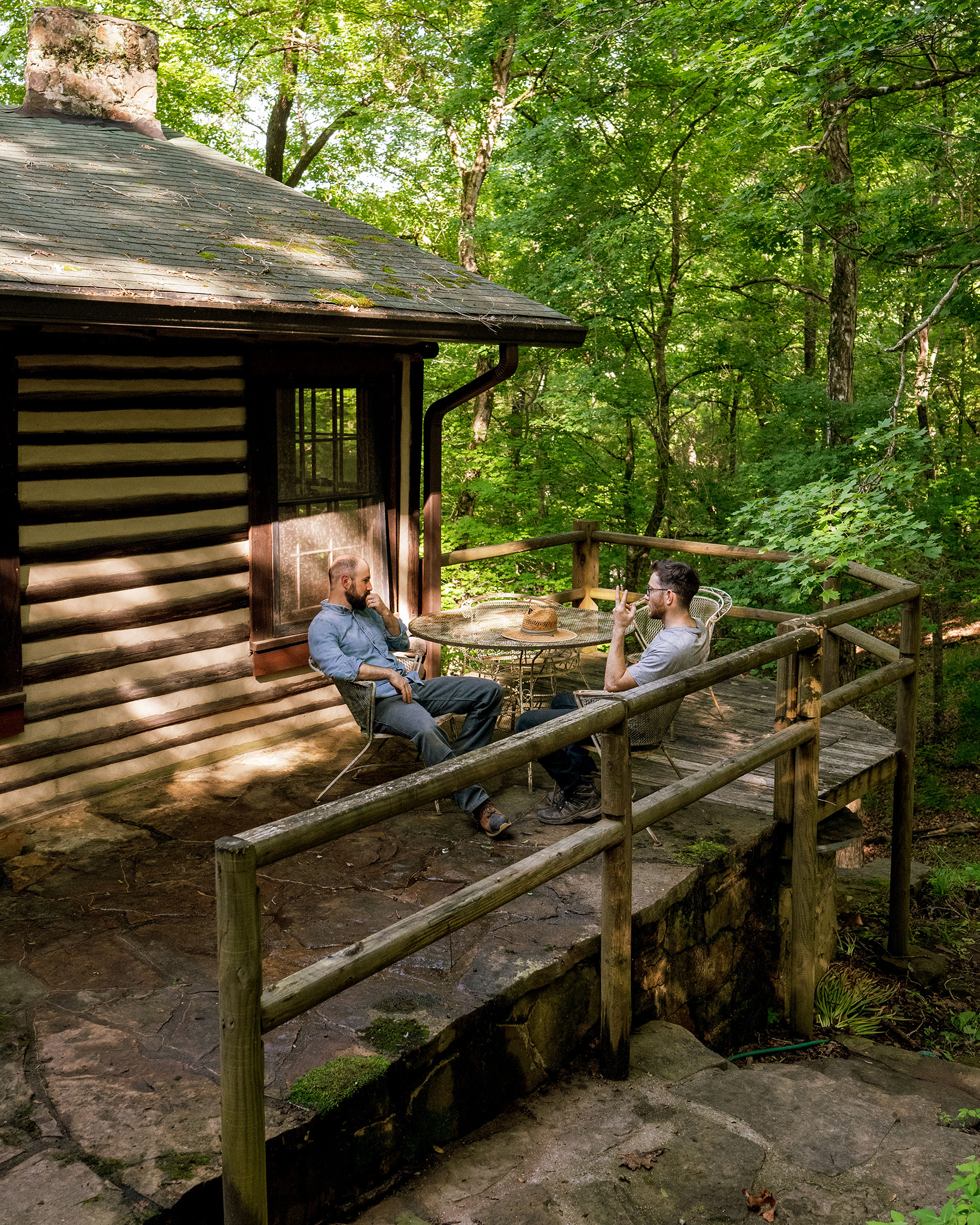 StickBuilt_Day1_018 _.5@0.5x Charlie and Will sit chatting around a table on the porch of a rustic cabin in the woods