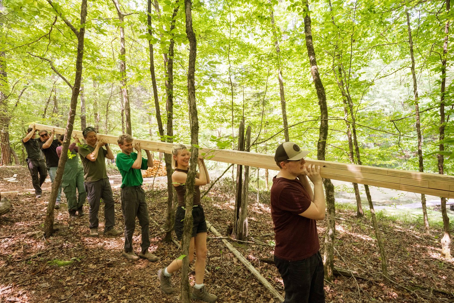 StickBuilt_Day3_218 Group of architect students and practitioners carrying a long piece of lumber together through the woods