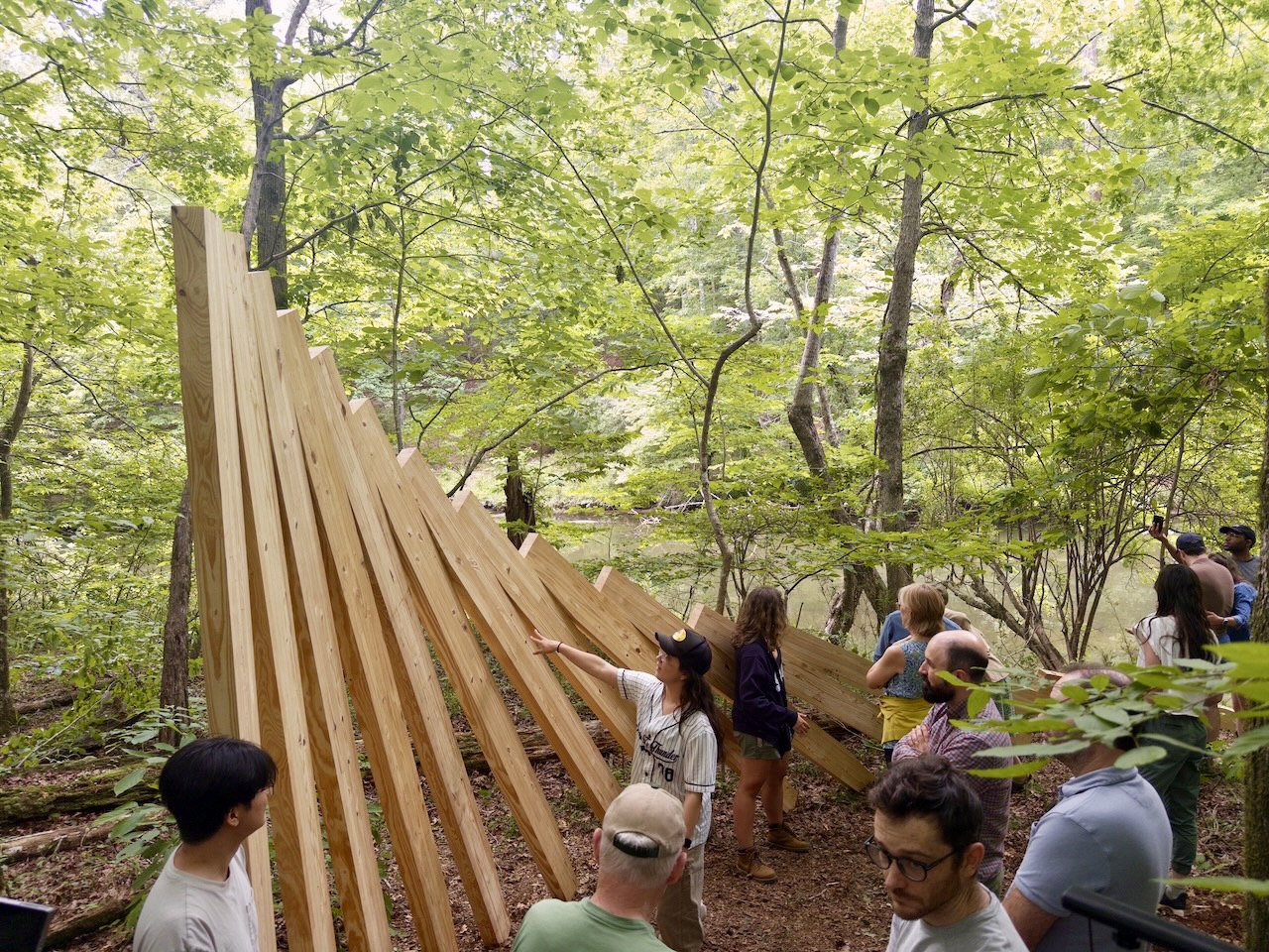 IMG_0432 2 Large pieces of lumber stacked on top of each other, cascading in a domino effect. The wood closest to the camera is standing upright, while the last piece of wood is laying on the ground. The structure is surrounded by trees and follows the riverline behind it.